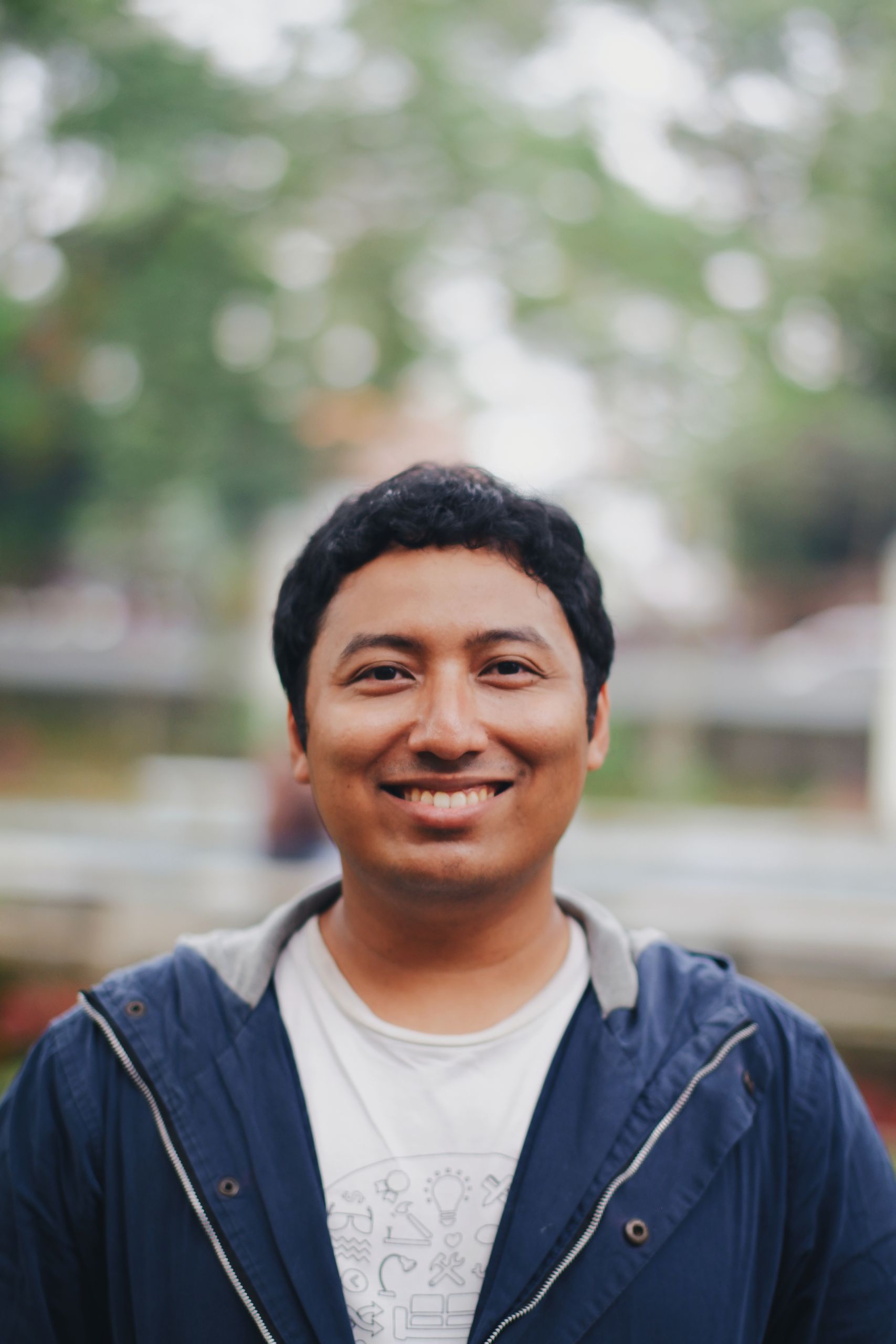 A cheerful man in a blue jacket smiles in an outdoor park setting.